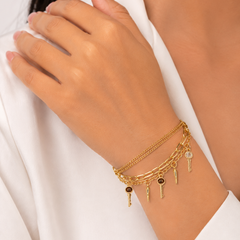 A close-up of a person's wrist wearing a delicate gold charm bracelet with key and chain charms, styled with a white top.