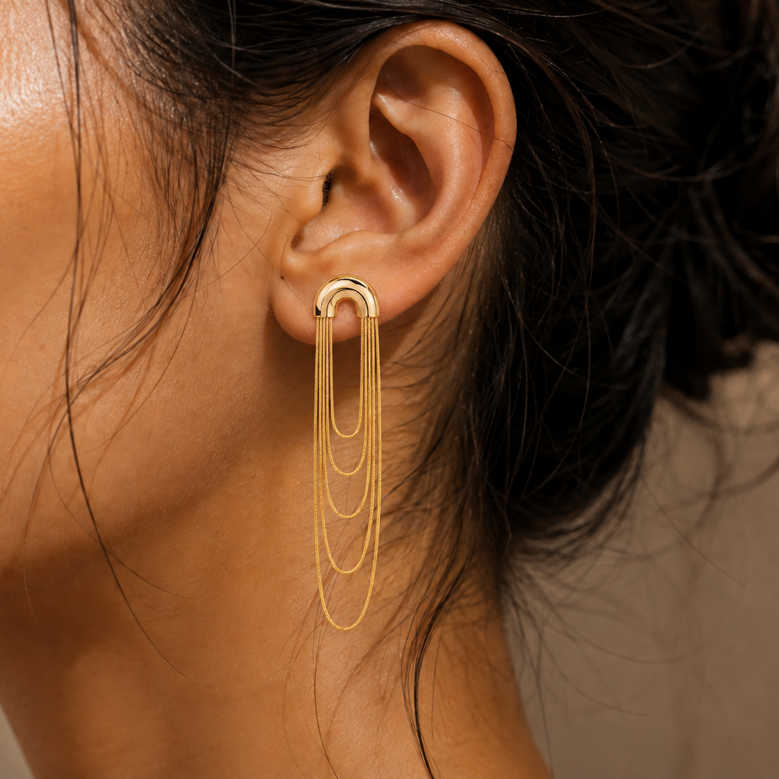 A close-up of a model's ear wearing a gold hoop earring with three cascading chain dangles against a neutral beige background.