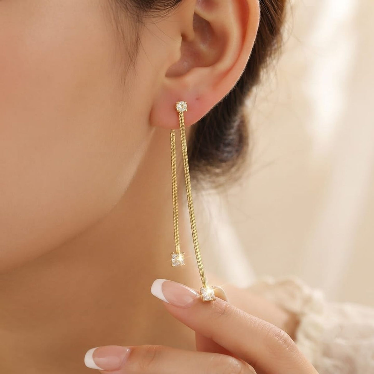 A close-up of a model's ear wearing a delicate gold dangle earring with a small clear gemstone at each end, while her fingers gently touch the lower dangling part.