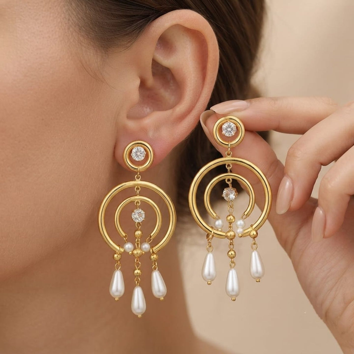 A close-up of a model wearing gold dangle earrings with white pearl drops and small clear stones, while a hand holds the matching earring against a soft beige background.