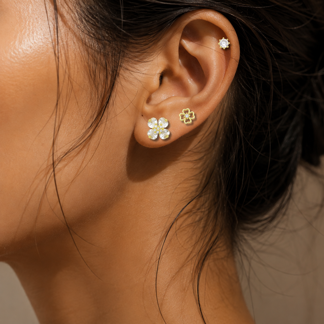 Close-up of a woman's ear wearing three small floral stud earrings in gold and white, with dark hair pulled back against a soft beige background.