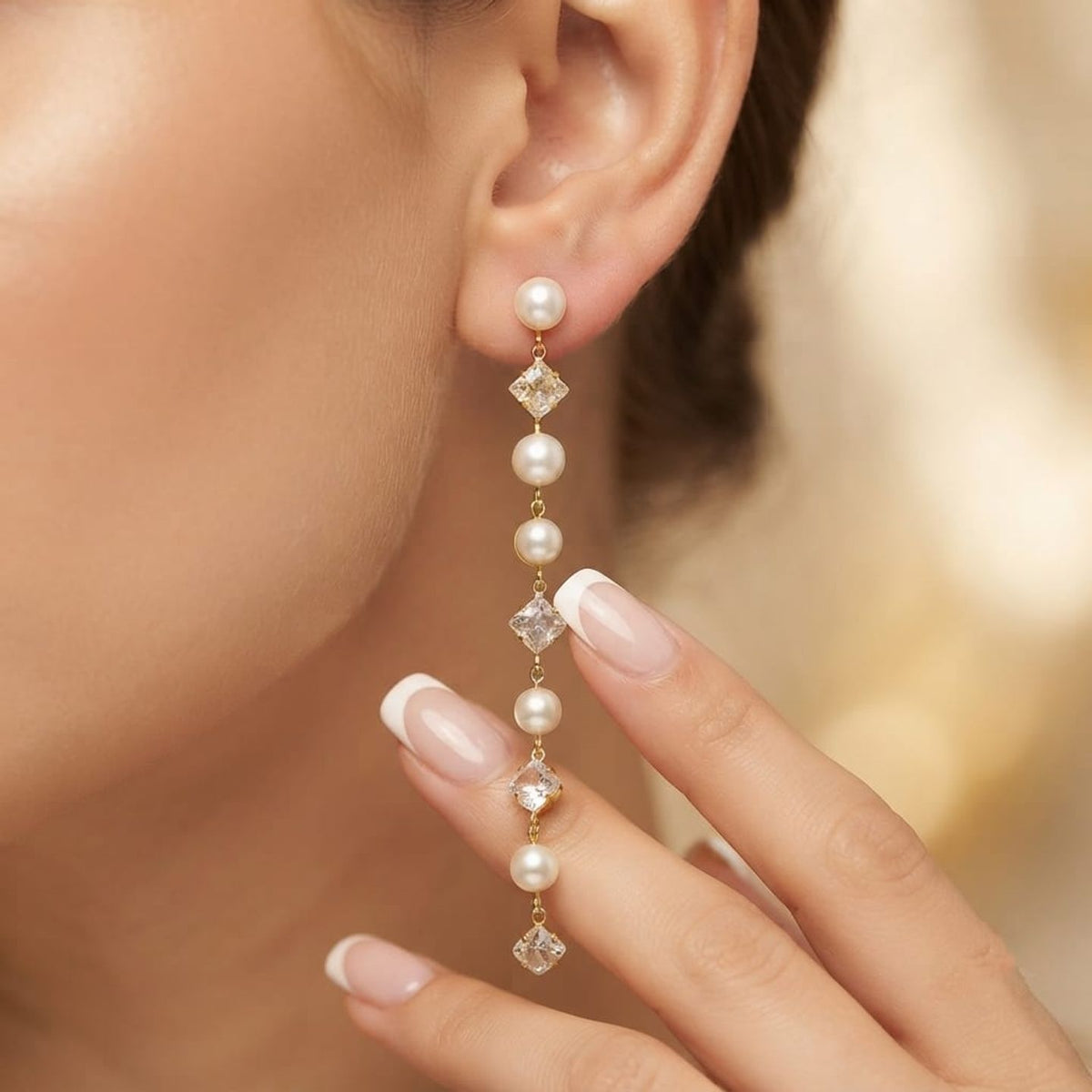 A close-up of a model's ear and hand wearing a long drop earring. The earring features a gold-tone setting with a series of white pearls and clear rhinestone-like diamond-shaped accents.
