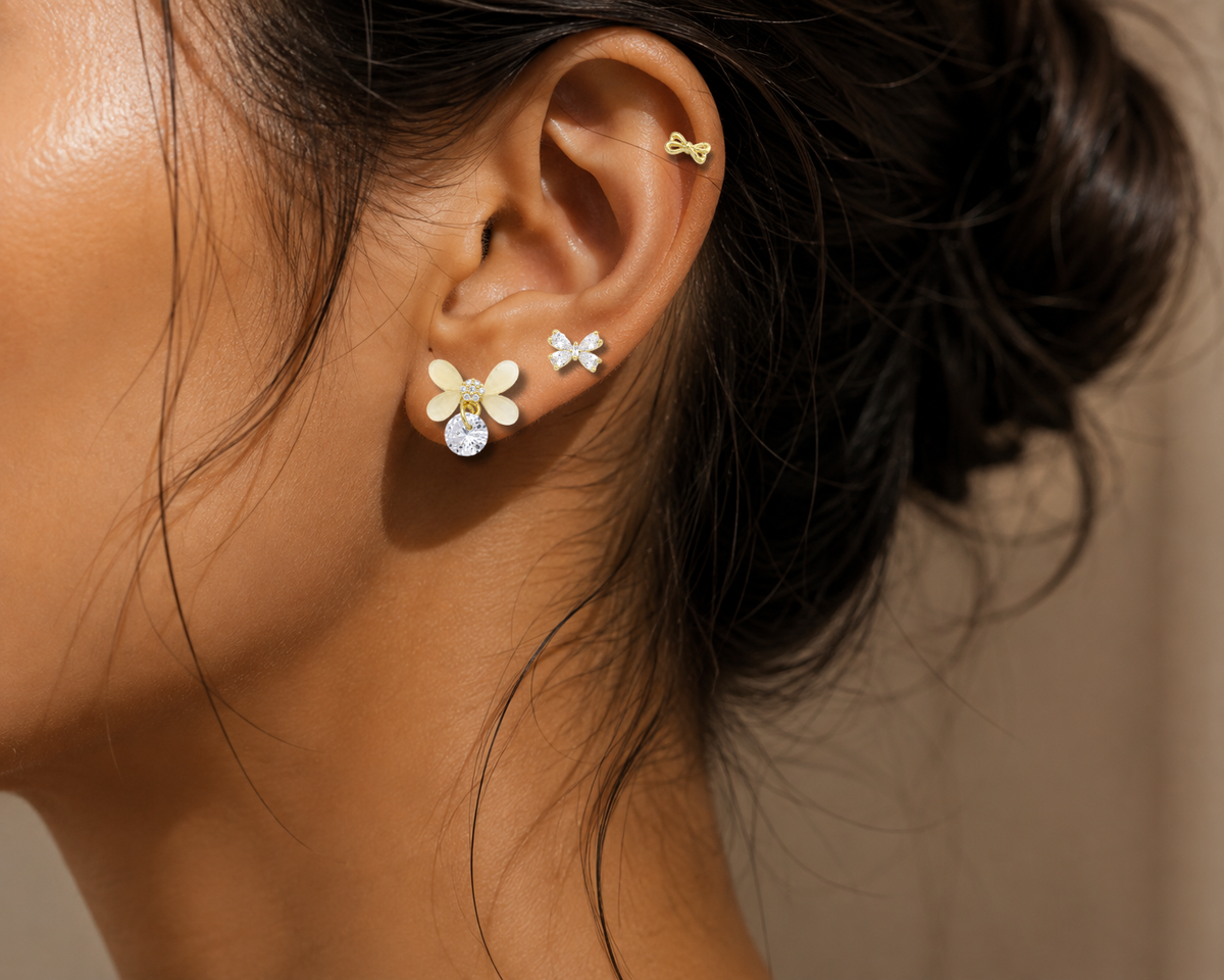 Close-up of a woman's ear wearing three small gold earrings: a bow stud, a flower stud with a clear rhinestone, and a butterfly stud, against a warm beige background.