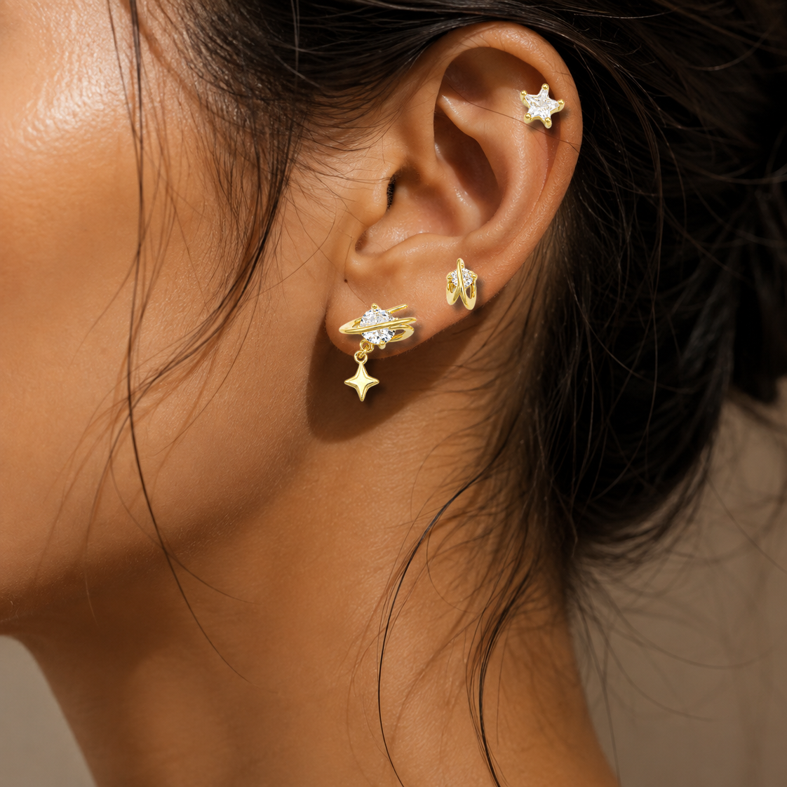 Close-up of a person's ear wearing multiple gold-toned ear piercings, including a star-shaped stud, a small crescent moon stud, and a larger crescent moon with a dangling star charm, against a soft beige background.