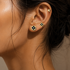 Close-up of a model's ear showcasing a stack of four gold ear piercings: a small star in the cartilage, a square stud with a black clover motif, a square stud with a white clover motif, and a flat gold hoop.