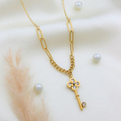 A gold chain necklace featuring a split design with a paperclip link section and a key-shaped pendant adorned with small blue and clear rhinestones, displayed on a soft cream fabric with decorative pearls and pampas grass.