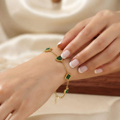 A delicate gold chain bracelet with green heart and diamond-shaped stones, shown worn on a wrist with manicured nails resting on the hand, set against a soft cream-colored background.