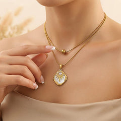 A close-up of a model wearing a double-layered gold necklace with a sunburst pendant featuring a white pearl center, styled with a beige top and soft neutral background.