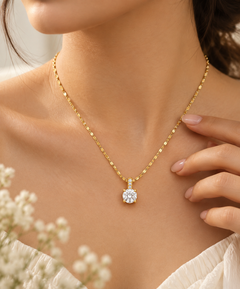 A close-up lifestyle shot of a model wearing a delicate gold chain necklace with a round-cut clear gemstone pendant. The necklace is displayed on the model's neck, with her hand resting near the chain, set against a soft, warm background with blurred white flowers in the foreground.