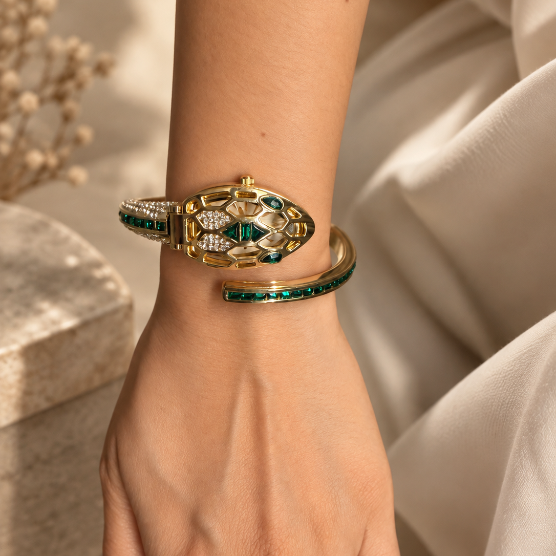 A gold-toned snake-head bangle bracelet with green emerald-cut stones and clear pavé accents, shown worn on a woman's wrist against a soft beige fabric background.