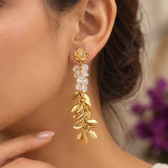 A close-up of a model's ear wearing a gold dangle earring with a floral stud, clear faceted crystal beads, and a leafy gold dangling charm.