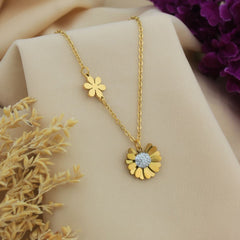 A gold-tone floral pendant necklace displayed on a beige fabric drape, featuring a small flower charm and a larger flower pendant with a clear rhinestone center, styled with dried flowers and purple blossoms.