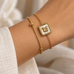 A close-up of a person's wrist wearing two delicate gold bracelets: one with a smooth bangle and bead, and one with a square white inlay featuring a gold butterfly design.