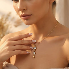 A close-up lifestyle shot of a woman wearing a delicate gold necklace with a multi-stone pendant. The necklace features a fine gold chain, a small round clear stone, a larger oval clear stone, a tiny gold bead, and a small gold heart.