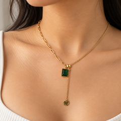 A gold Y-necklace with a rectangular green gemstone pendant and a dangling gold coin charm, shown worn on a model's neck.