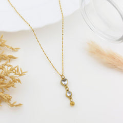 A delicate gold chain necklace with a vertical drop pendant featuring three clear round gemstones in gold settings and a small dangling gold bead, styled on a white surface with dried grass and a glass rim.