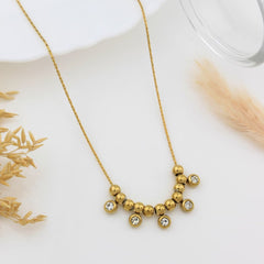 A gold necklace with a curved bar of small gold beads and five bezel-set clear rhinestones, displayed on a white surface with dried decorative grass.