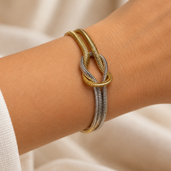 A close-up of a person's wrist wearing a three-band bangle bracelet with a central knot design. The bands are textured snake-style metal in gold and silver tones, set against a soft beige fabric background.