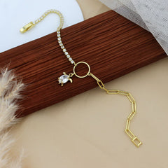 A gold-toned chain bracelet with a row of clear rhinestones, a small clear crystal charm, and a circular ring connector, displayed on a dark wood block with beige fabric and pampas grass props.