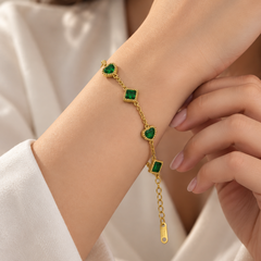 A gold chain bracelet with three green heart and square gemstone charms, shown worn on a woman's wrist.