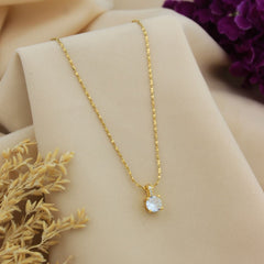 A gold chain necklace with a heart-shaped clear gemstone pendant, displayed on a beige fabric jewelry stand with dried grass and purple flowers in the background.