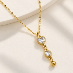 A gold dangle necklace featuring a heart-shaped clear crystal, a round clear crystal, and a small gold bead, displayed on a soft cream background.