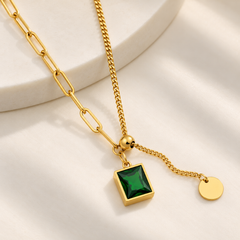 Gold chain necklace with a square green gemstone pendant and a small gold disc charm, styled on a white marble pedestal against a soft white background.