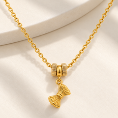 A gold chain necklace with a bow-shaped pendant and two textured cylindrical beads, displayed on a soft cream-colored fabric background.