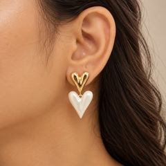 A close-up of a model's ear wearing a gold heart stud earring with a dangling white heart charm.