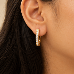 A close-up of a person's ear wearing a gold huggie hoop earring set with clear pavé gemstones, against a soft beige background.