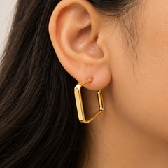 A close-up of a model's ear wearing a gold geometric hoop earring with a square open frame design.