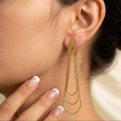 A close-up of a model wearing a long, geometric gold dangle earring with three nested teardrop loops. The model's ear, neck, and hand with a French manicure are visible against a soft, light beige background.