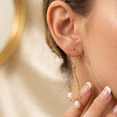 A close-up of a woman's ear wearing a gold hoop earring with dangling twisted gold chains and white pearl drops, against a soft cream background.