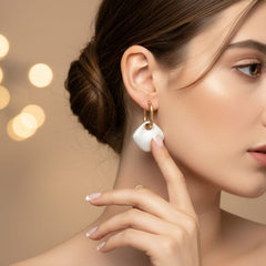 A close-up lifestyle shot of a woman wearing a gold hoop earring with a white, organic-shaped drop pendant. The background is a soft, warm beige with blurred bokeh lights.