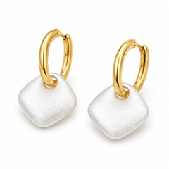 A pair of gold huggie hoop earrings with a textured, square-shaped white pendant dangling from each hoop, shown on a clean white background.