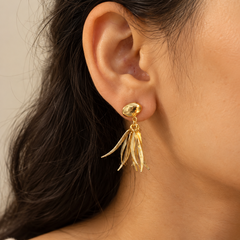A close-up of a model's ear wearing a gold dangle earring with a textured oval top and three layered feather-like drops.