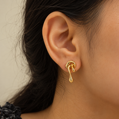 A close-up of a person's ear wearing a small gold hoop earring with a dangling teardrop-shaped drop, shown against a soft beige background.